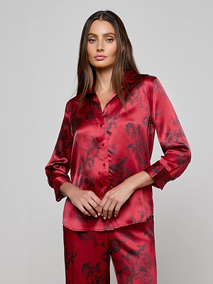 A woman models the Dani Silk Blouse by L'AGENCE—a burgundy floral silk top—standing against a plain gray background, looking at the camera with her hands gently clasped in front of her.