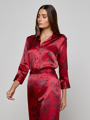 A woman wears the Dani Silk Blouse by L'AGENCE, a red satin floral print top paired with matching trousers, standing against a plain light background—an elegant, stylish choice for any wardrobe.