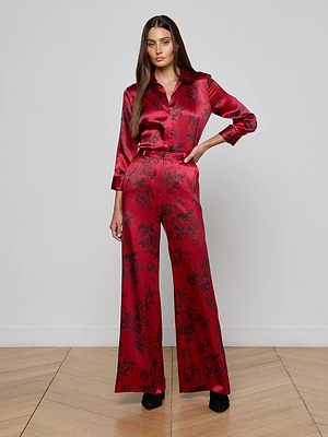 A woman with long brown hair and a neutral expression wears the Dani Silk Blouse by L'AGENCE in burgundy, styled with a shiny red pantsuit featuring a black floral pattern and paired with black shoes against a plain light background.
