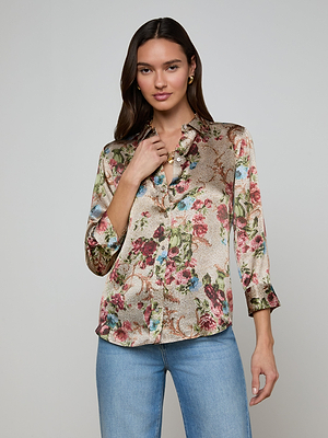 A woman with long brown hair wears a shiny Dani Silk Blouse in a floral tapestry print with rolled sleeves and blue jeans, standing against a plain light-colored background while looking at the camera and touching her necklace.