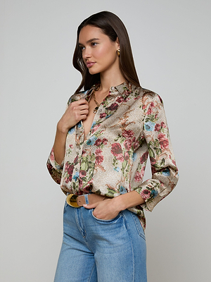 A woman with long brown hair wears the Dani Silk Blouse—a shiny floral-print button-down—paired with blue jeans, a brown belt, gold earrings, and a gold necklace, standing against a plain light background.