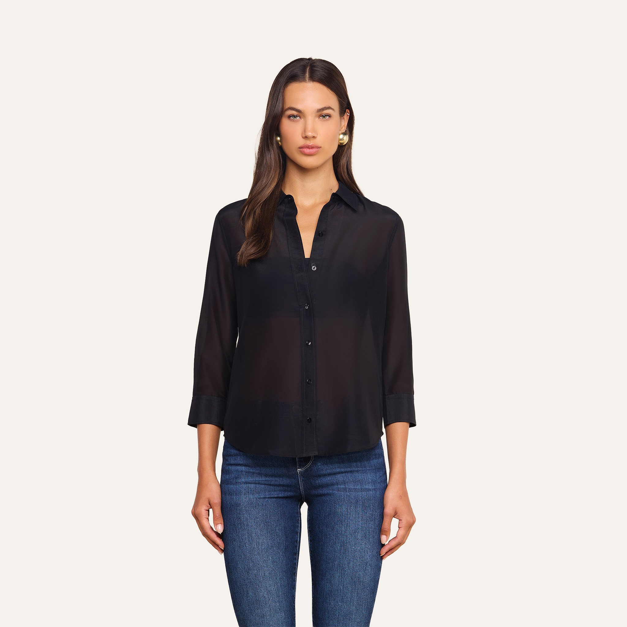 A woman with long brown hair wears the Dani Cotton-Silk Blouse—a black button-down top with 3/4 sleeves—paired with blue jeans, standing against a plain, light background.