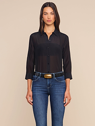 A woman stands facing forward against a plain light background, wearing the Dani Cotton-Silk Blouse—a sheer black button-down with 3/4 sleeves—paired with blue jeans and a black belt featuring a gold buckle.