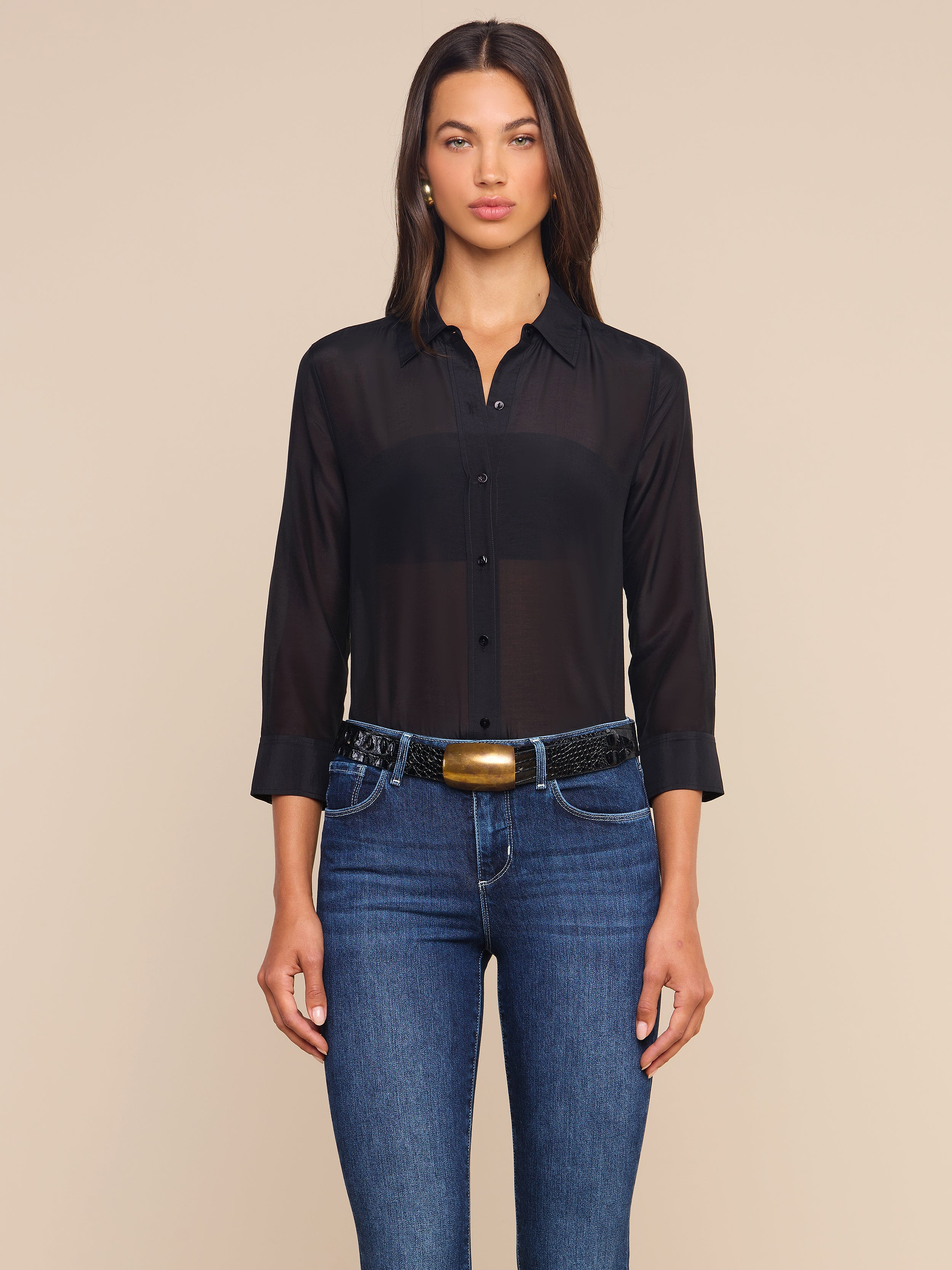 A woman stands facing forward against a plain light background, wearing the Dani Cotton-Silk Blouse—a sheer black button-down with 3/4 sleeves—paired with blue jeans and a black belt featuring a gold buckle.