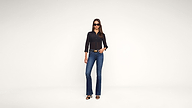 A woman stands confidently in a black top, blue jeans, sunglasses, and heels against a plain white background, with one hand in her pocket.