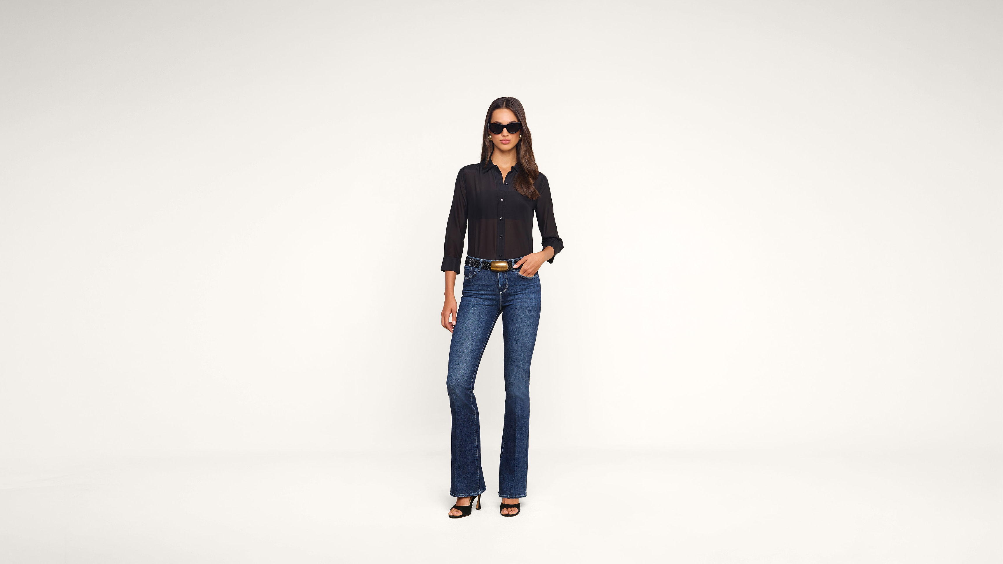 A woman stands confidently in a black top, blue jeans, sunglasses, and heels against a plain white background, with one hand in her pocket.
