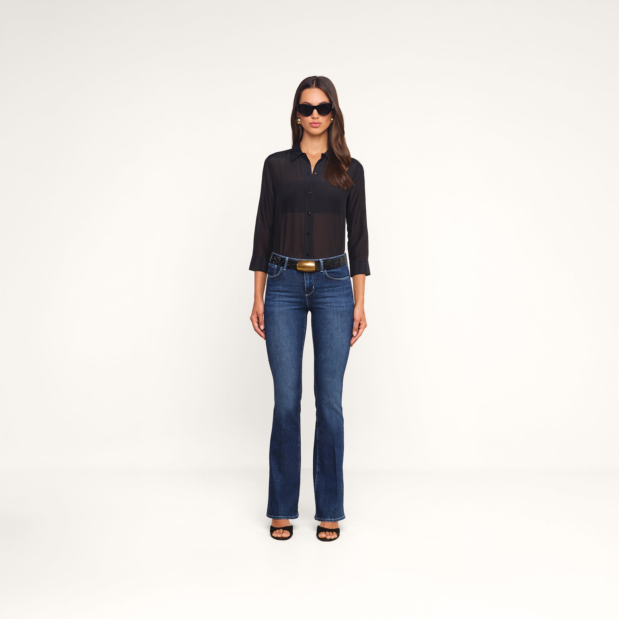 A woman wearing sunglasses, the Dani Cotton-Silk Blouse—a sheer black button-down with 3/4 sleeves—paired with blue jeans, a belt featuring a large buckle, and black open-toe heels stands facing forward against a plain white background.
