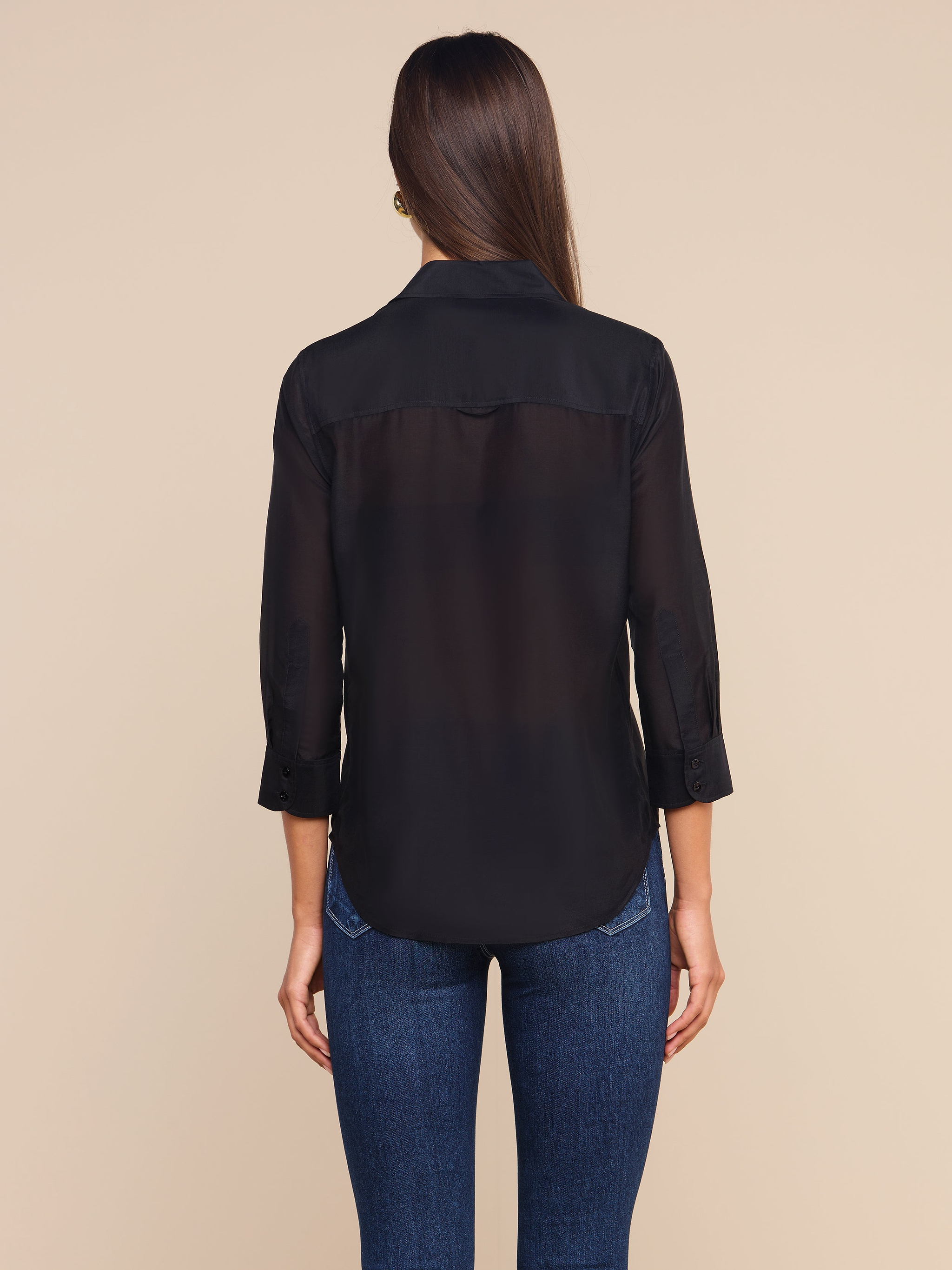 A woman stands facing away, wearing the Dani Cotton-Silk Blouse—a sheer black button-down with 3/4 sleeves—paired with blue jeans against a plain light background.