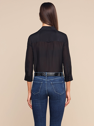 A woman with long brown hair, wearing a black Dani Cotton-Silk Blouse and blue jeans with a black belt, stands facing away from the camera against a plain beige background.