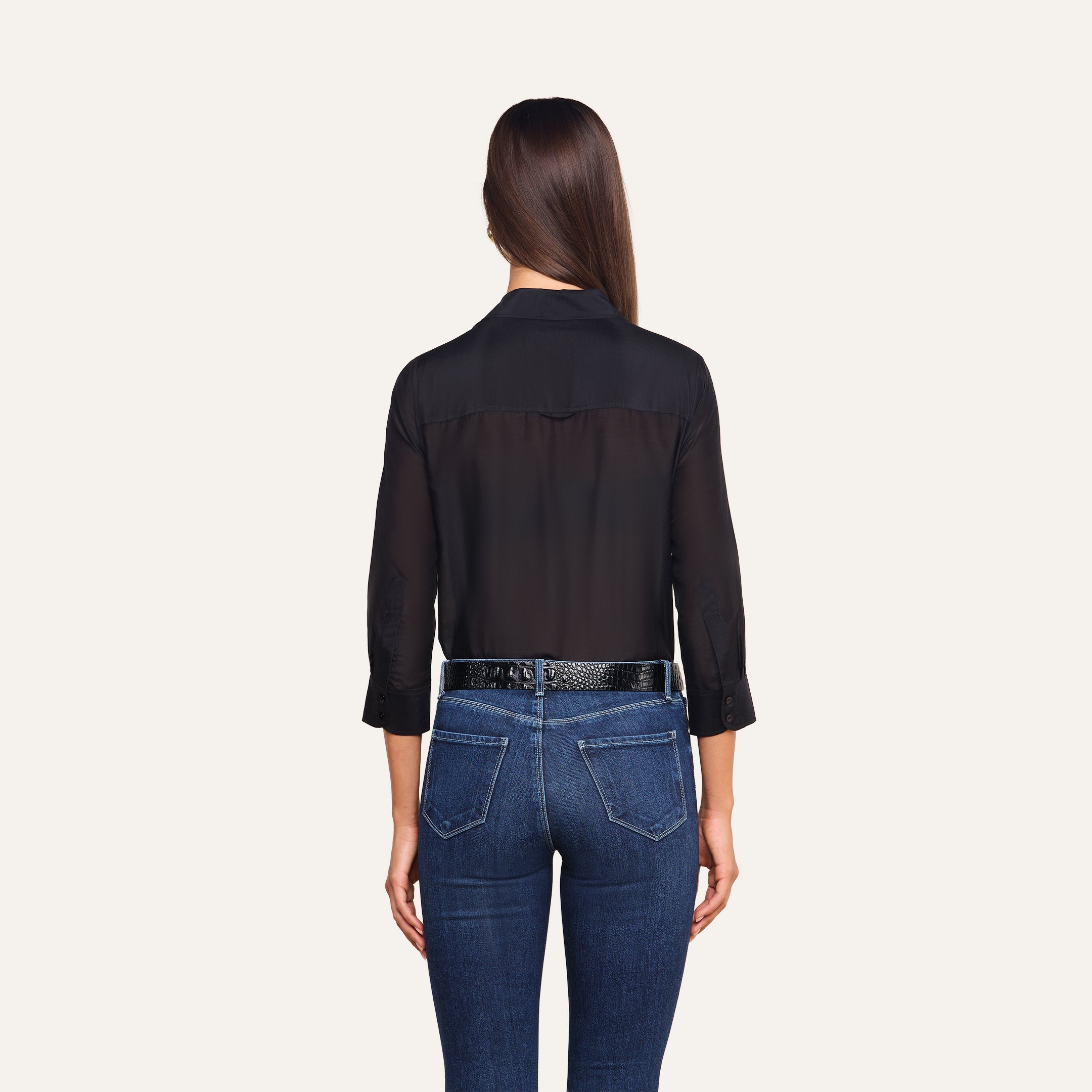 A woman with long brown hair, wearing the Dani Cotton-Silk Blouse—a sheer black button-down top with 3/4 sleeves—paired with a black belt and blue jeans, stands facing away from the camera against a plain background.