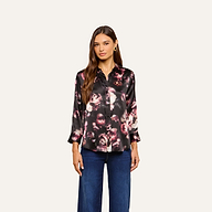 A woman models the Dani Silk Blouse—a long-sleeve, black silk button-down top with pink floral patterns—paired with blue jeans and posed against a plain light background.