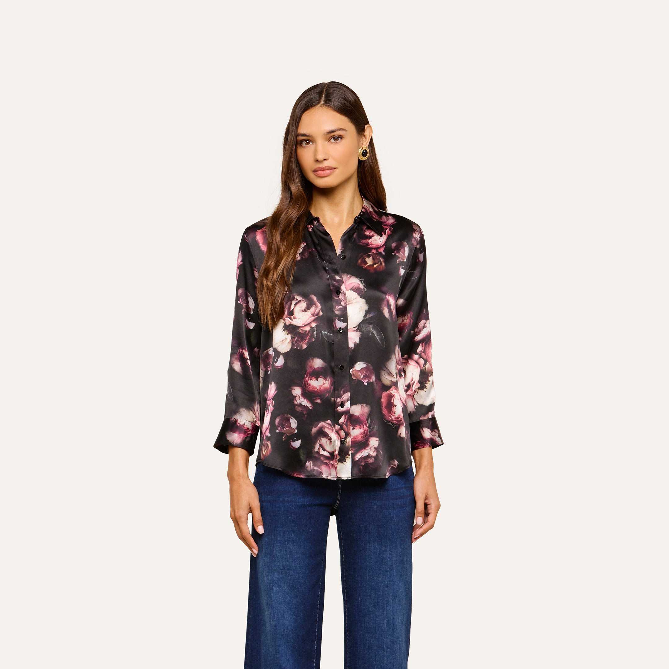 A woman models the Dani Silk Blouse—a long-sleeve, black silk button-down top with pink floral patterns—paired with blue jeans and posed against a plain light background.