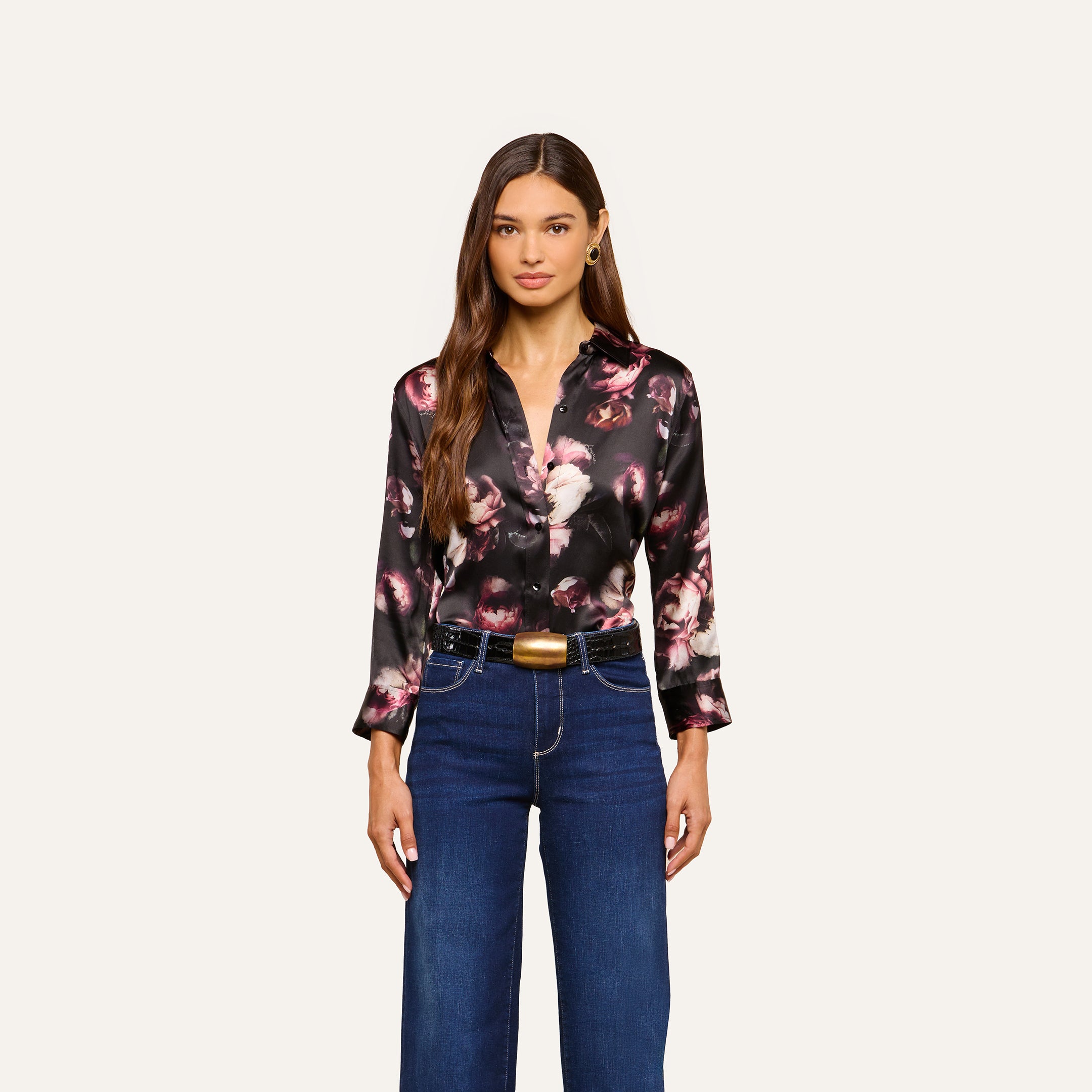 A woman wearing the Dani Silk Blouse— a black floral silk button-down— and high-waisted blue jeans stands against a plain light background, looking forward with her hands by her sides.