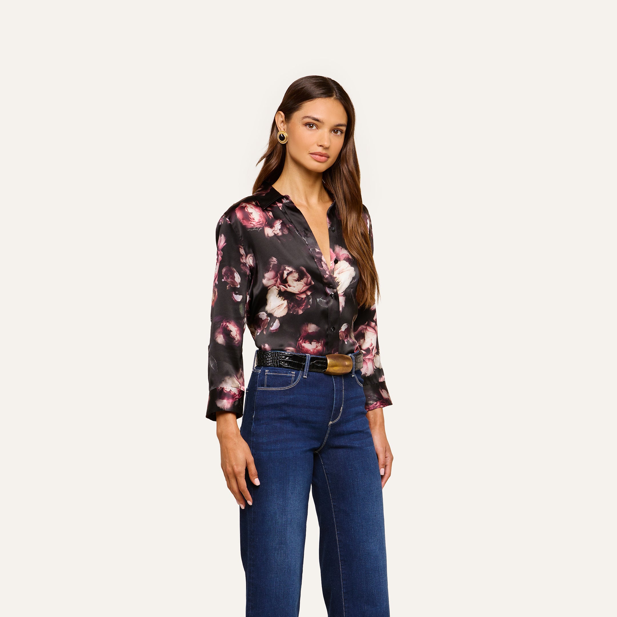 A woman with long brown hair and a neutral expression stands against a plain background wearing the Dani Silk Blouse—a black silk charmeuse top featuring a floral pattern—paired with dark blue jeans, a belt with a gold buckle, and gold hoop earrings.