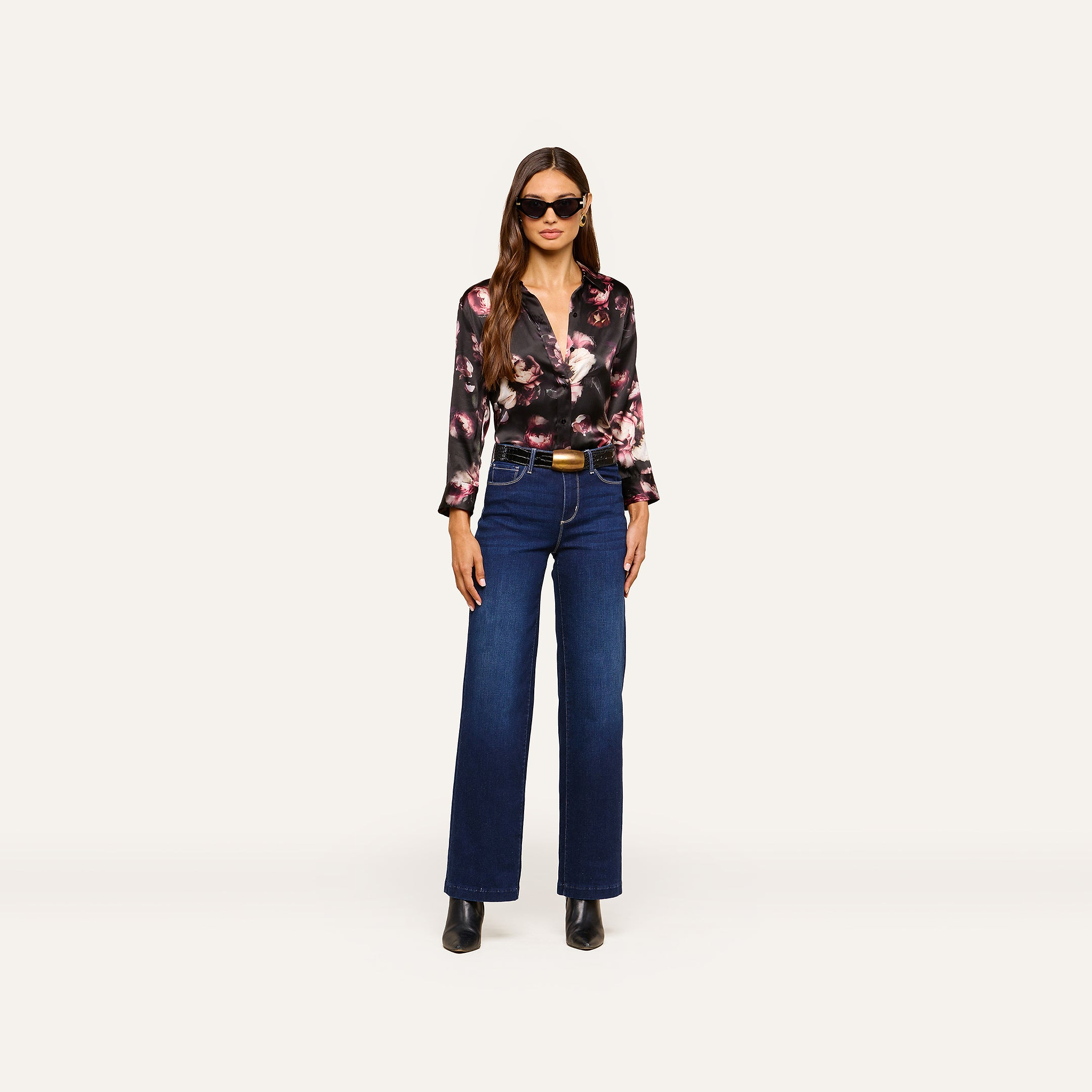 A woman with long brown hair and a confident posture stands against a plain background wearing sunglasses, a Dani Silk Blouse in a floral print, dark blue high-waisted wide-leg jeans, a brown belt, and black boots.