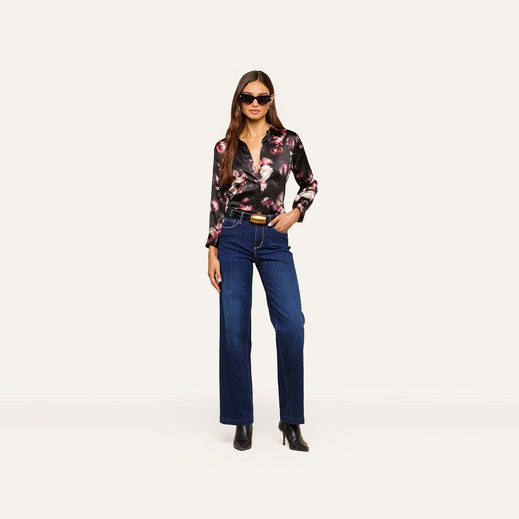 A woman stands confidently against a plain light background, wearing sunglasses, a black floral Dani Silk Blouse in luxurious silk charmeuse, paired with dark blue high-waisted jeans and black heeled boots.