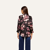 A woman stands facing away, wearing the Dani Silk Blouse in a pink floral print with blue jeans against a plain light background.
