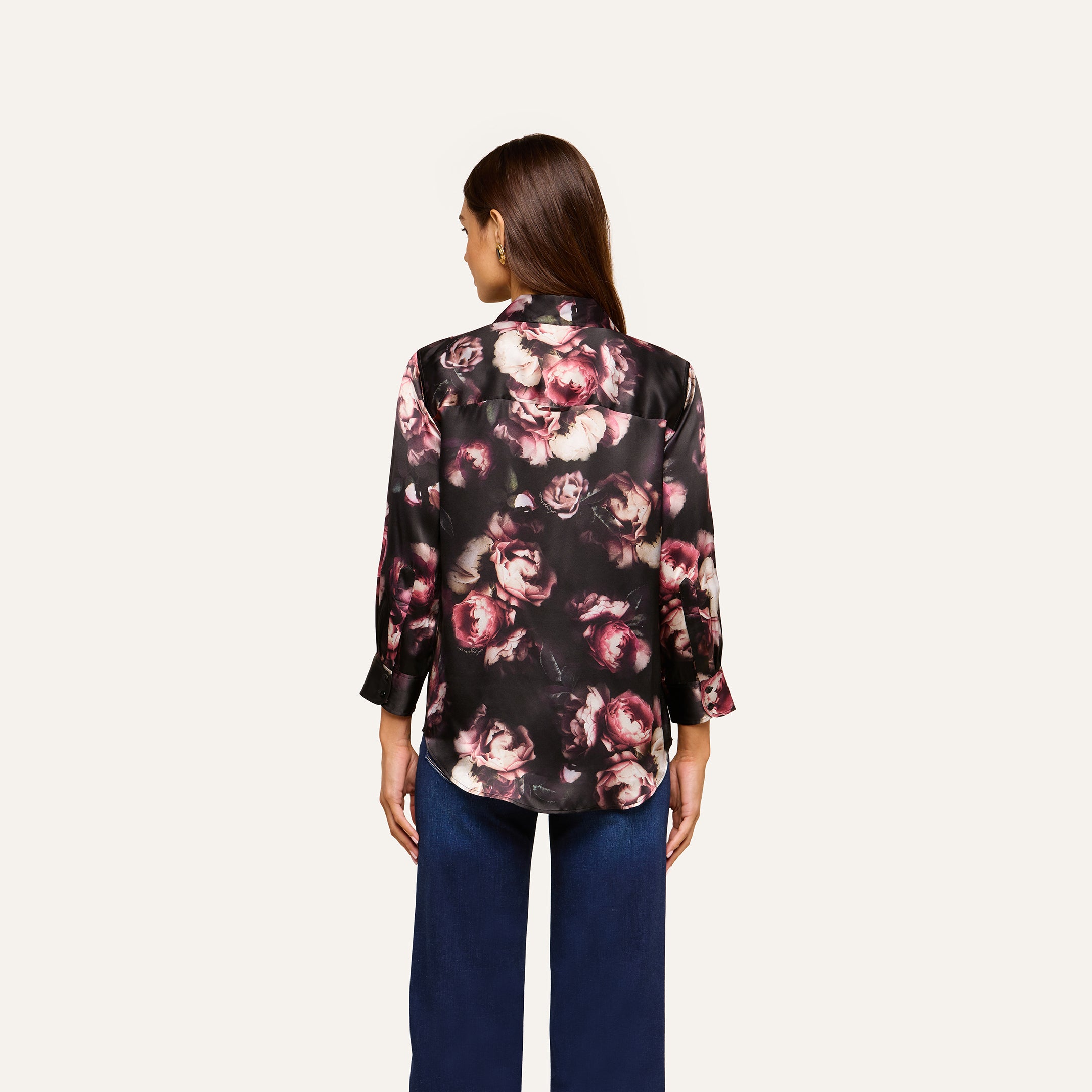 A woman stands facing away, wearing the Dani Silk Blouse in a pink floral print with blue jeans against a plain light background.