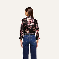 A woman stands with her back to the camera, wearing the Dani Silk Blouse in black floral silk charmeuse adorned with pink and cream roses, styled with high-waisted blue jeans. The background is plain and light-colored.