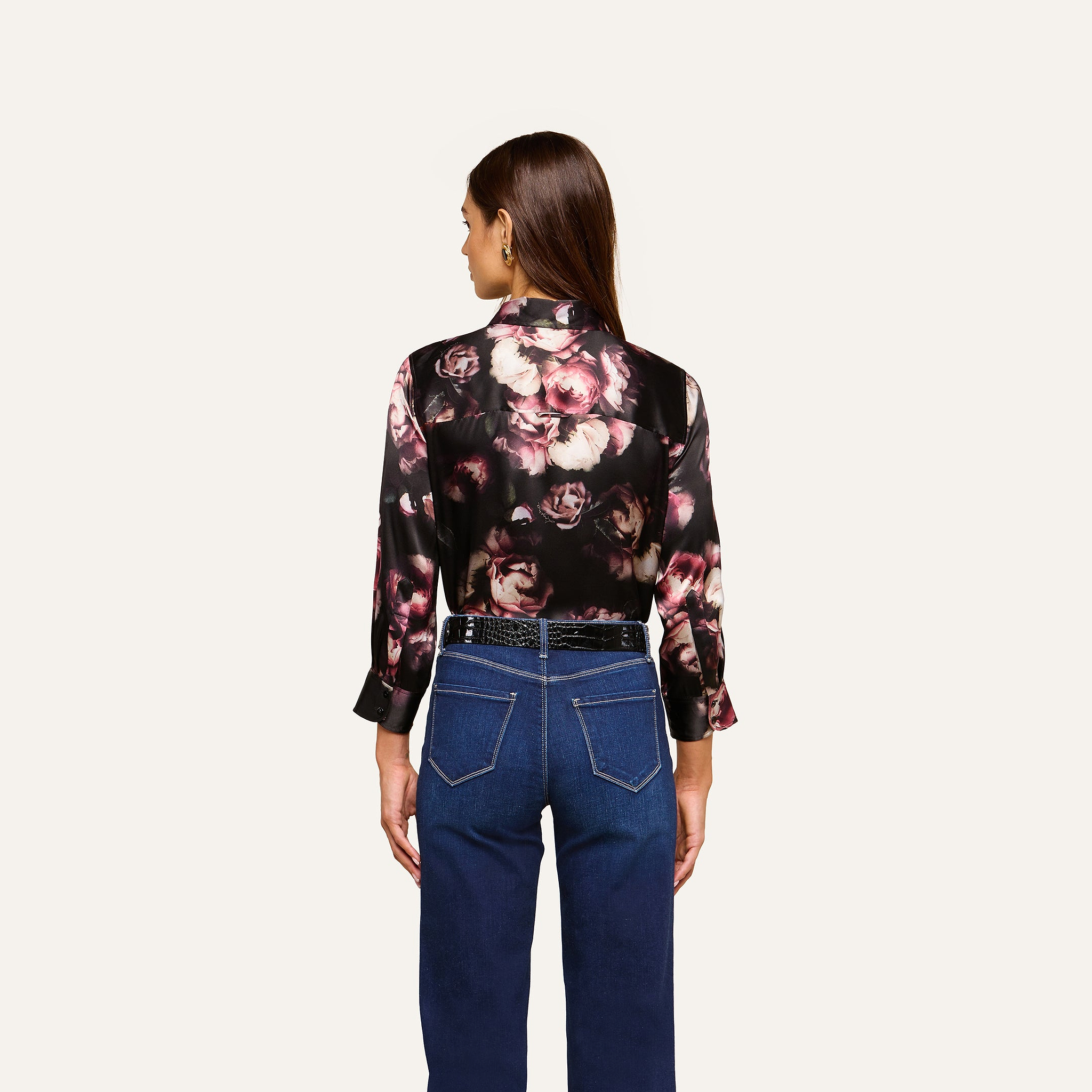 A woman stands with her back to the camera, wearing the Dani Silk Blouse in black floral silk charmeuse adorned with pink and cream roses, styled with high-waisted blue jeans. The background is plain and light-colored.