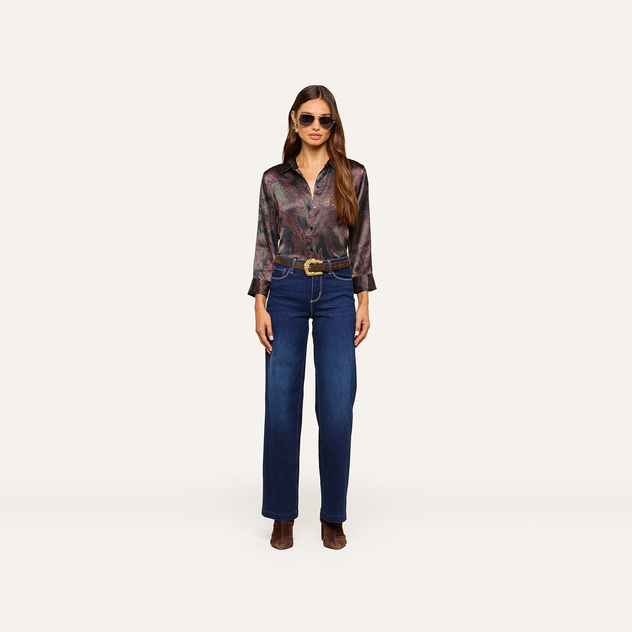 Woman wearing sunglasses and the Dani Silk Blouse—a paisley silk button-down with three-quarter sleeves—paired with a wide brown belt, high-waisted blue jeans, and brown heeled boots, standing against a plain white background.