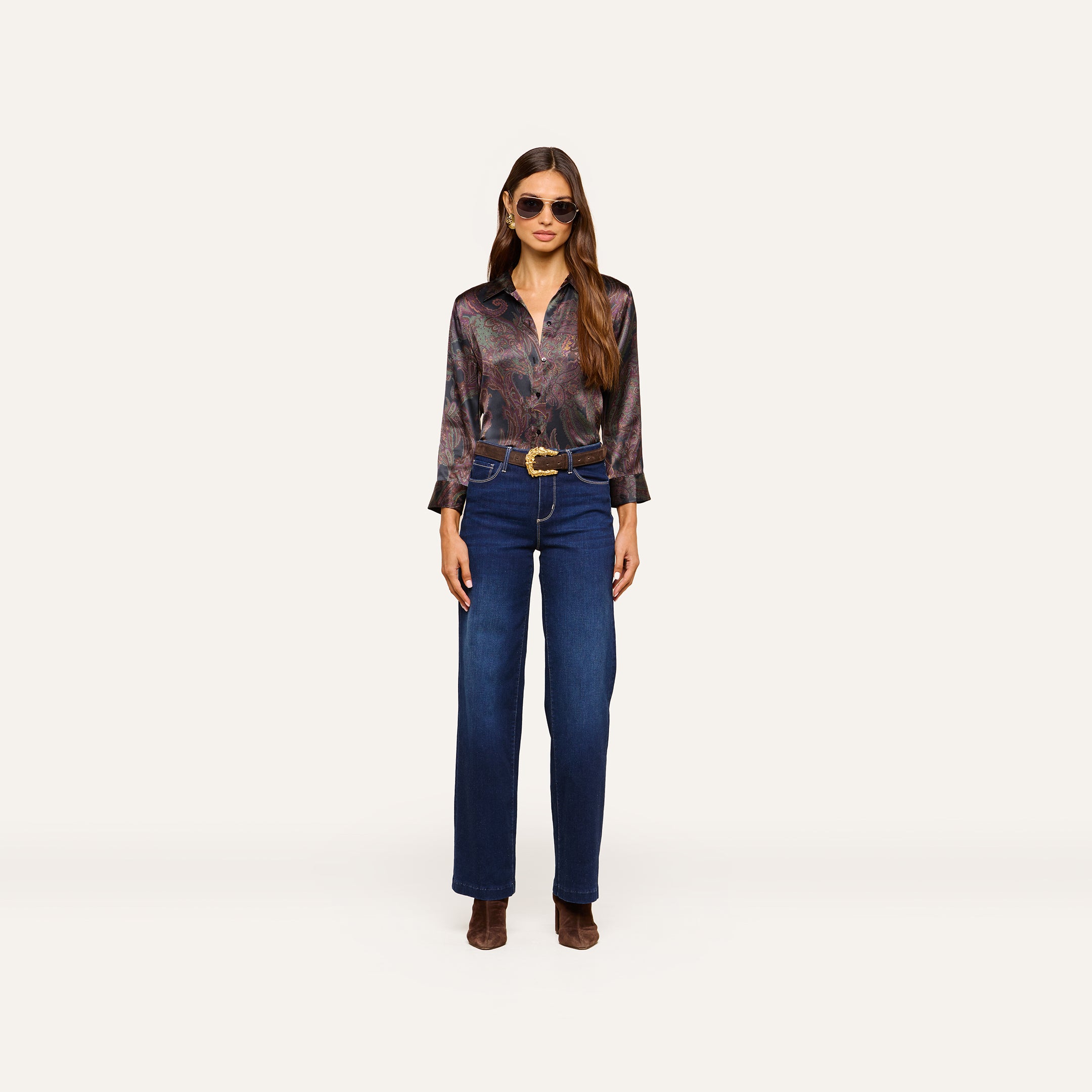 Woman wearing sunglasses and the Dani Silk Blouse—a paisley silk button-down with three-quarter sleeves—paired with a wide brown belt, high-waisted blue jeans, and brown heeled boots, standing against a plain white background.