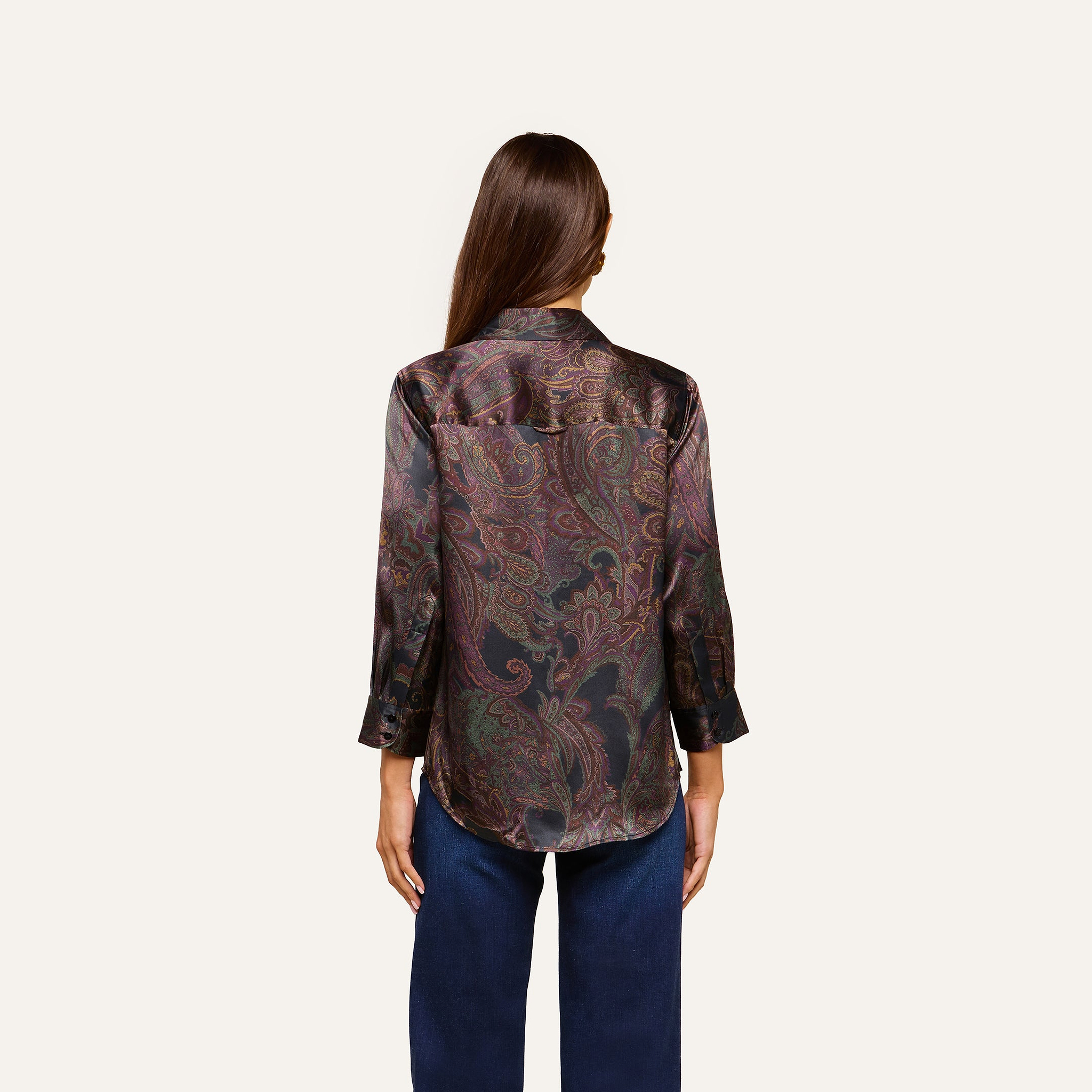 A woman with long brown hair, seen from the back, wears the Dani Silk Blouse—a dark paisley top with three-quarter sleeves—paired with blue jeans against a plain light background.
