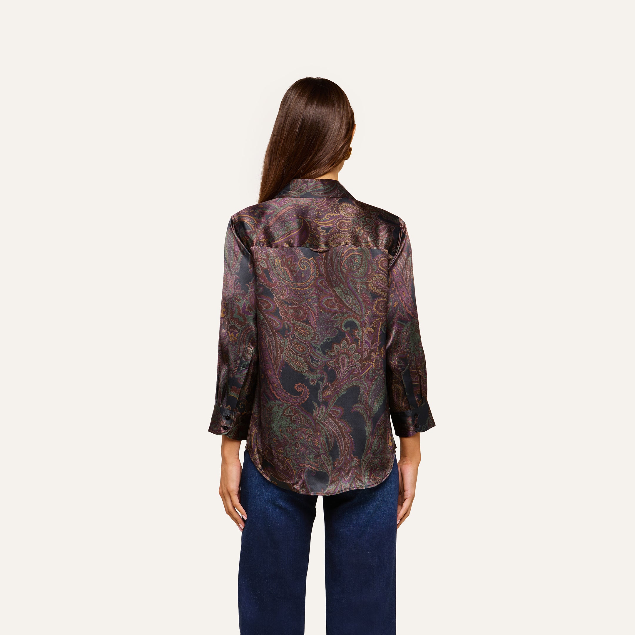 A woman with long brown hair, seen from the back, wears the Dani Silk Blouse—a dark paisley top with three-quarter sleeves—paired with blue jeans against a plain light background.