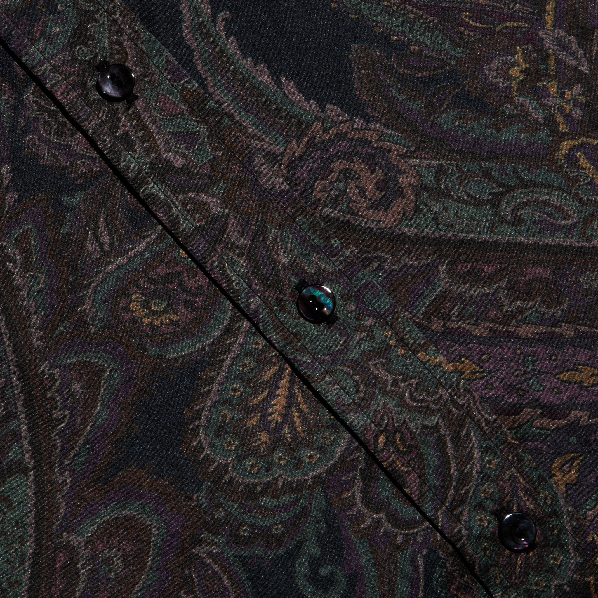 Close-up of the Dani Silk Blouse’s paisley-patterned silk fabric in dark colors, highlighting a diagonal seam with black buttons.