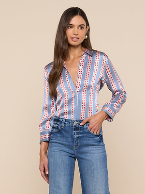 A woman stands against a beige background wearing the Dani Silk Blouse, a long-sleeve button-down with red, white, and blue vertical stripes. She pairs it with high-waisted blue jeans, her long brown hair loose and one hand in her pocket.