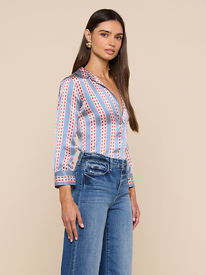A woman with long brown hair wears the Dani Silk Blouse, a blue, white, and red vertically striped button-down, paired with blue jeans. She stands against a beige background, looking slightly to the side.
