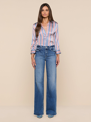 A woman with long brown hair faces forward in a striped Dani Silk Blouse with red and blue lines and high-waisted wide-leg blue jeans, standing against a plain beige background.