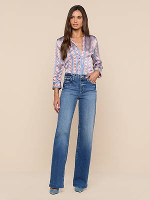 A woman with long brown hair and a neutral expression stands against a beige background, wearing the Dani Silk Blouse—featuring red, white, and blue vertical stripes—with high-waisted blue flare jeans and blue pointed-toe heels.