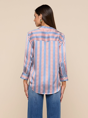 A woman with straight brown hair wears the Dani Silk Blouse, a button-down featuring vertical blue, white, and red-orange stripes with dots. She’s accessorized with gold hoop earrings and pairs the blouse with blue jeans.