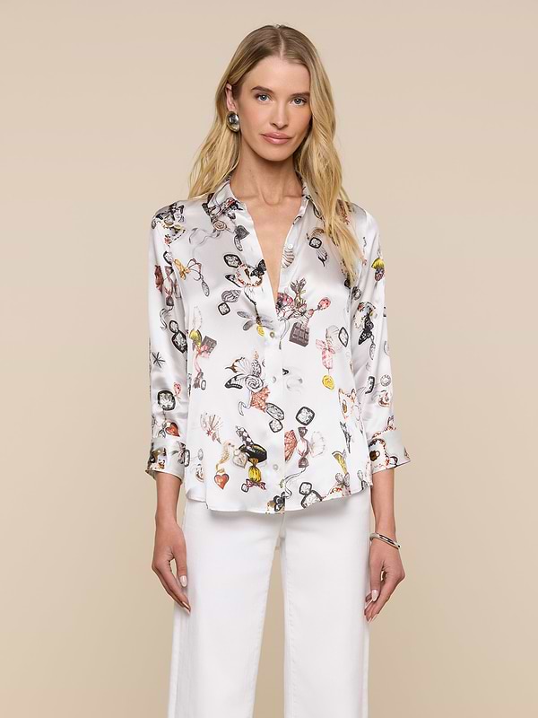 A woman with long blonde hair models the Dani Silk Blouse, featuring a white butterfly print, paired with white pants. She stands against a plain light background and looks at the camera with a slight smile.