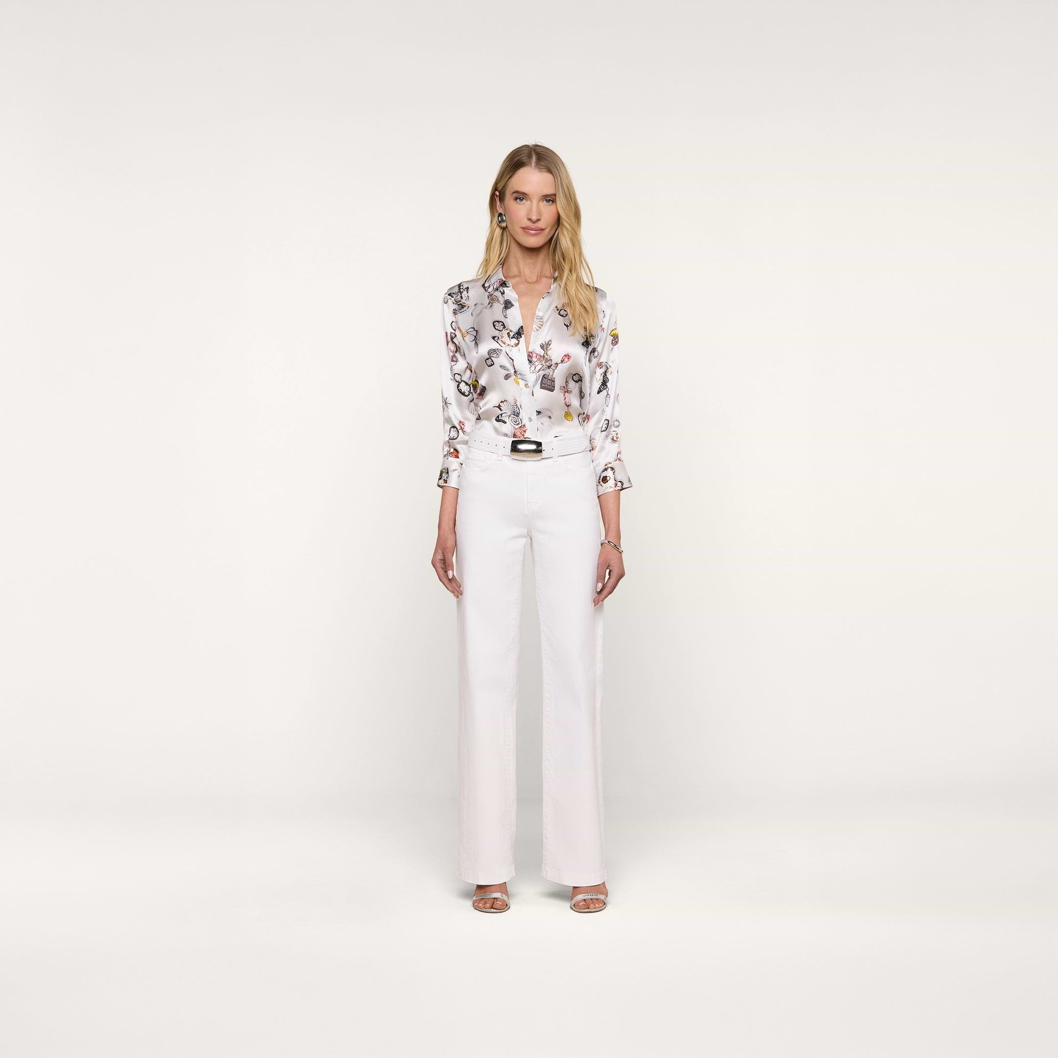 A woman stands facing forward against a plain white background, wearing the Dani Silk Blouse with a butterfly print, tucked into high-waisted white trousers and paired with heeled sandals.