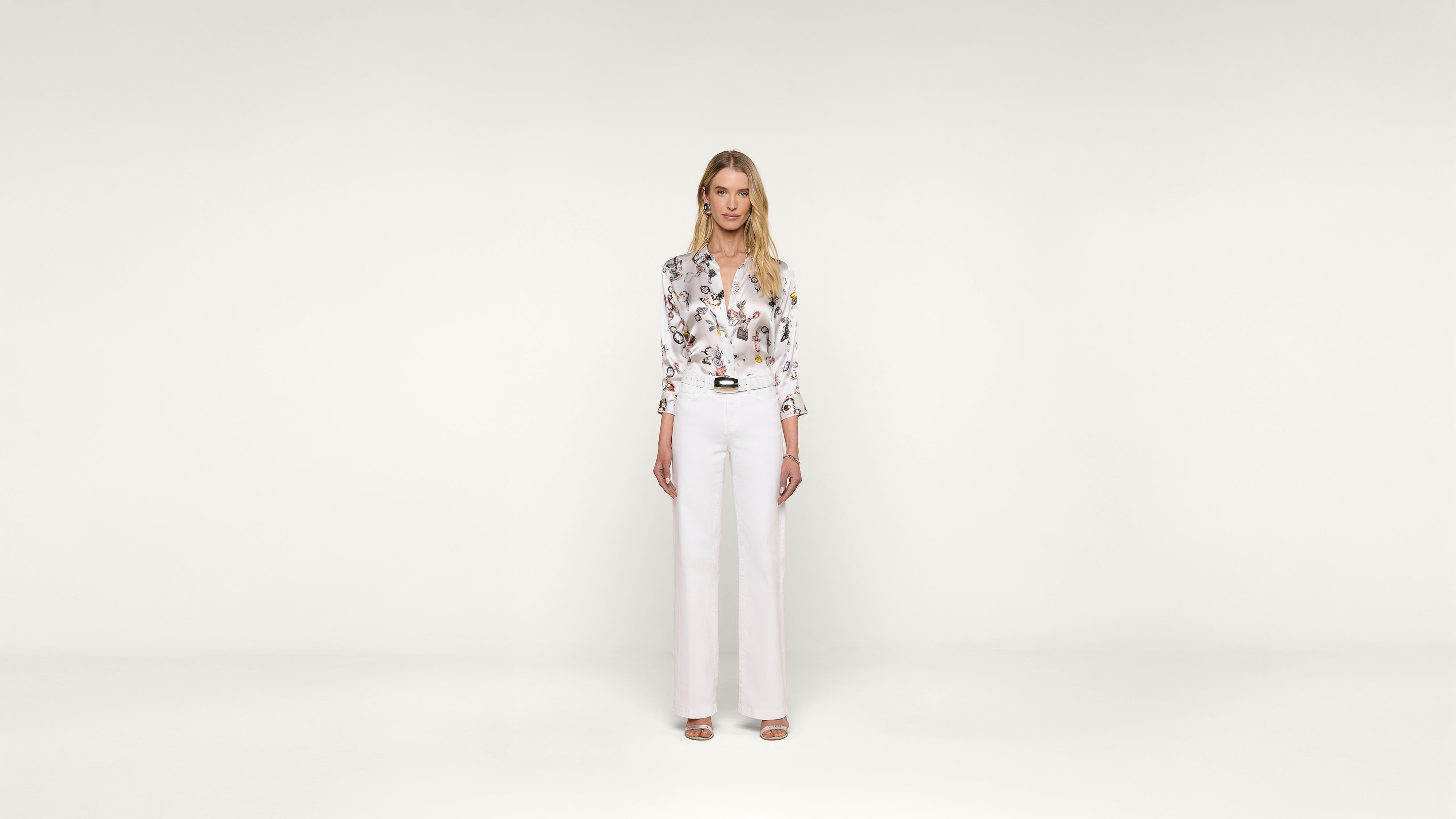 A woman stands facing forward, wearing a floral blouse tucked into high-waisted white pants and heels, against a plain white background.