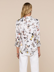 A woman stands facing away, wearing the Dani Silk Blouse—a white silk button-down adorned with a colorful butterfly print—and white pants, set against a plain light background.