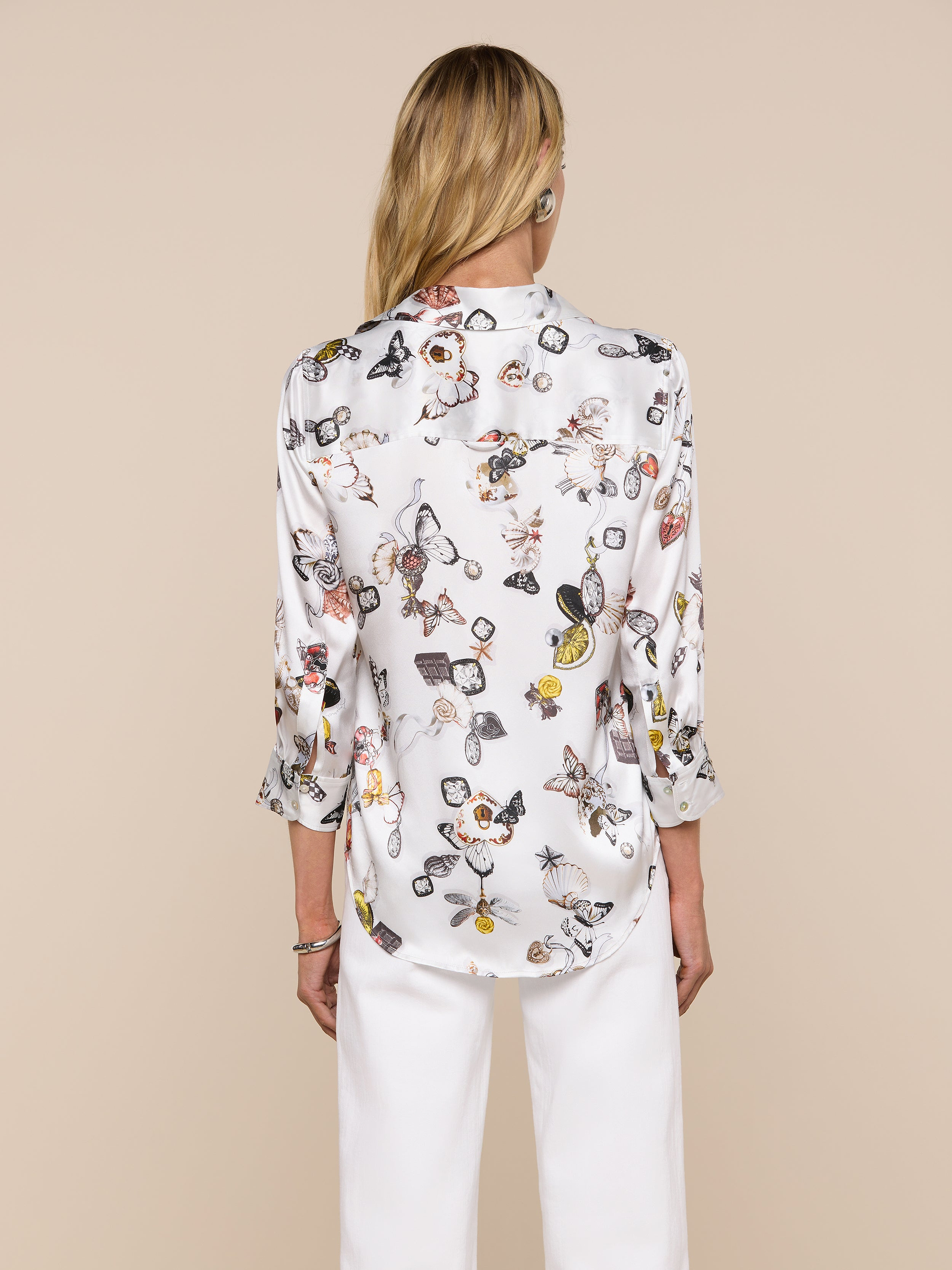 A woman stands facing away, wearing the Dani Silk Blouse—a white silk button-down adorned with a colorful butterfly print—and white pants, set against a plain light background.