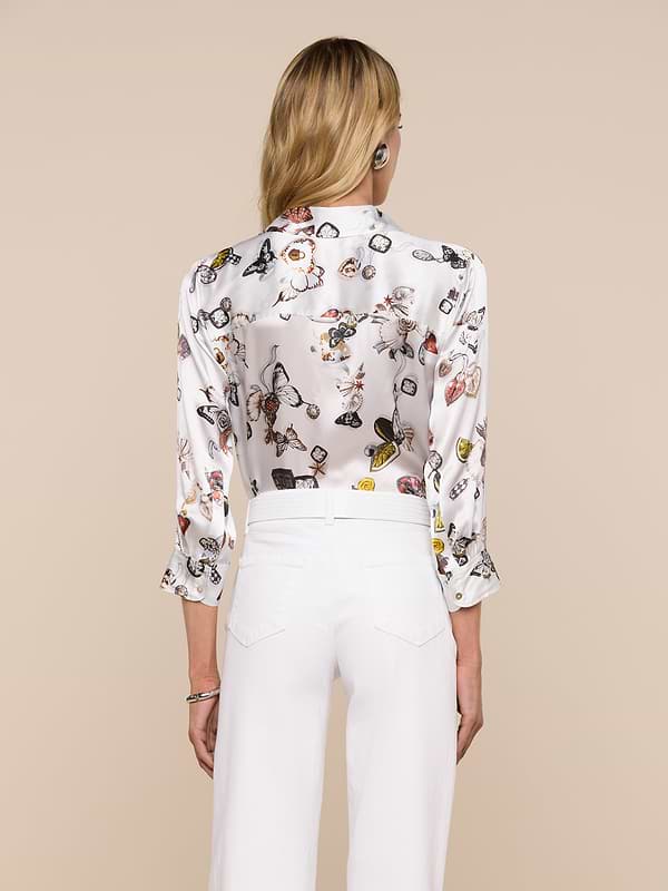 A woman with long blonde hair, dressed in the Dani Silk Blouse featuring a colorful print and paired with white pants, is shown from the back against a plain light background.