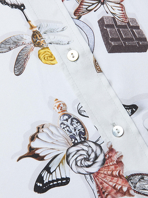 A close-up of the Dani Silk Blouse, a button-down shirt with white buttons, center seam, and a delicate pattern featuring butterflies, seashells, a yellow rose, an ornate bottle, and a piece of chocolate.
