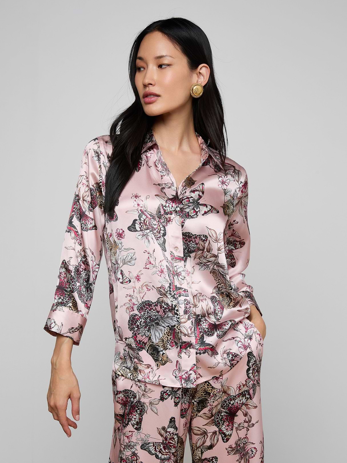 A woman wearing the Dani Silk Blouse in a shiny, light pink silk fabric with a butterfly and floral pattern poses with one hand in her pocket against a plain light background.