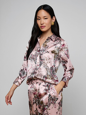 A woman with long dark hair wears the shiny pink Dani Silk Blouse and matching butterfly jungle print pants. She stands against a plain background, looking confidently at the camera with one hand in her pocket.