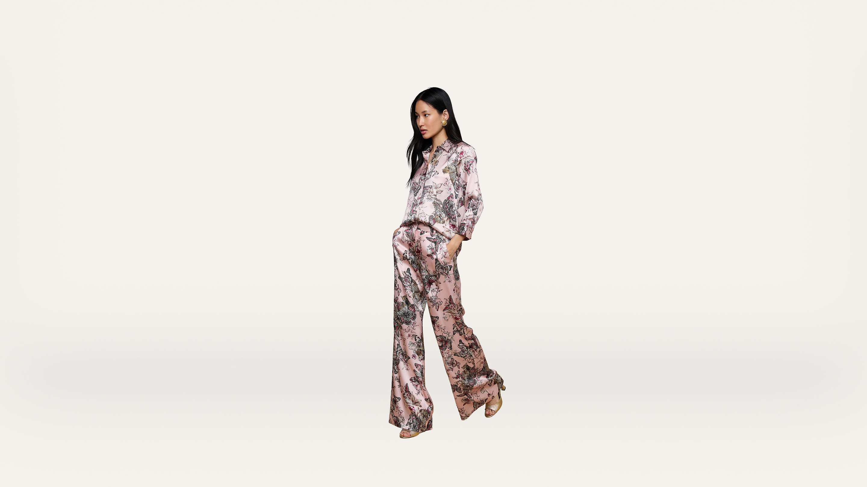 A woman stands against a plain white background, wearing a matching light pink floral blouse and wide-leg pants with her hands in her pockets.
