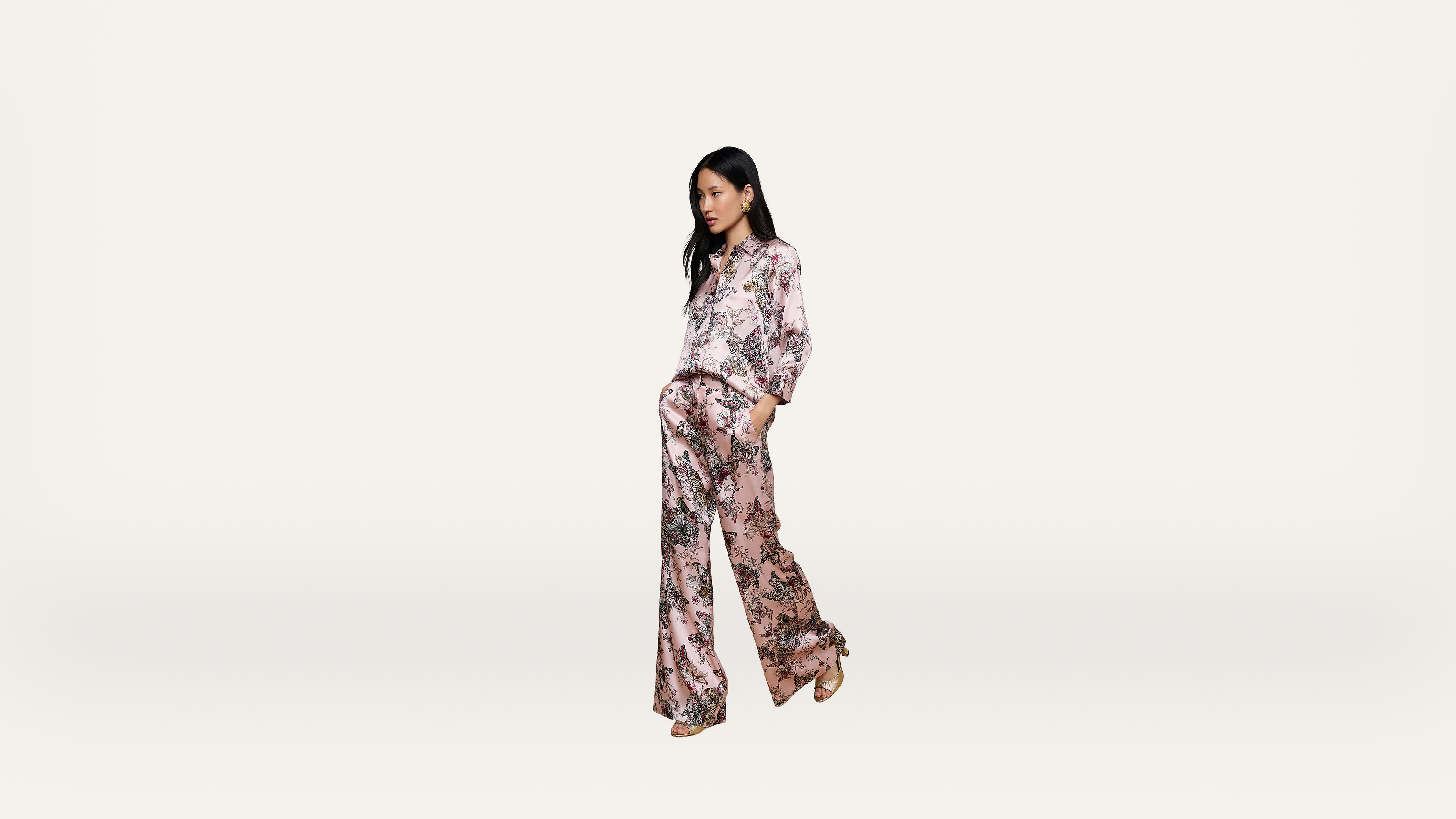 A woman stands against a plain white background, wearing a matching light pink floral blouse and wide-leg pants with her hands in her pockets.