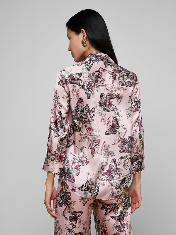 A woman with long dark hair, seen from the back, wears the Dani Silk Blouse featuring a pink butterfly jungle print, standing against a plain light background.