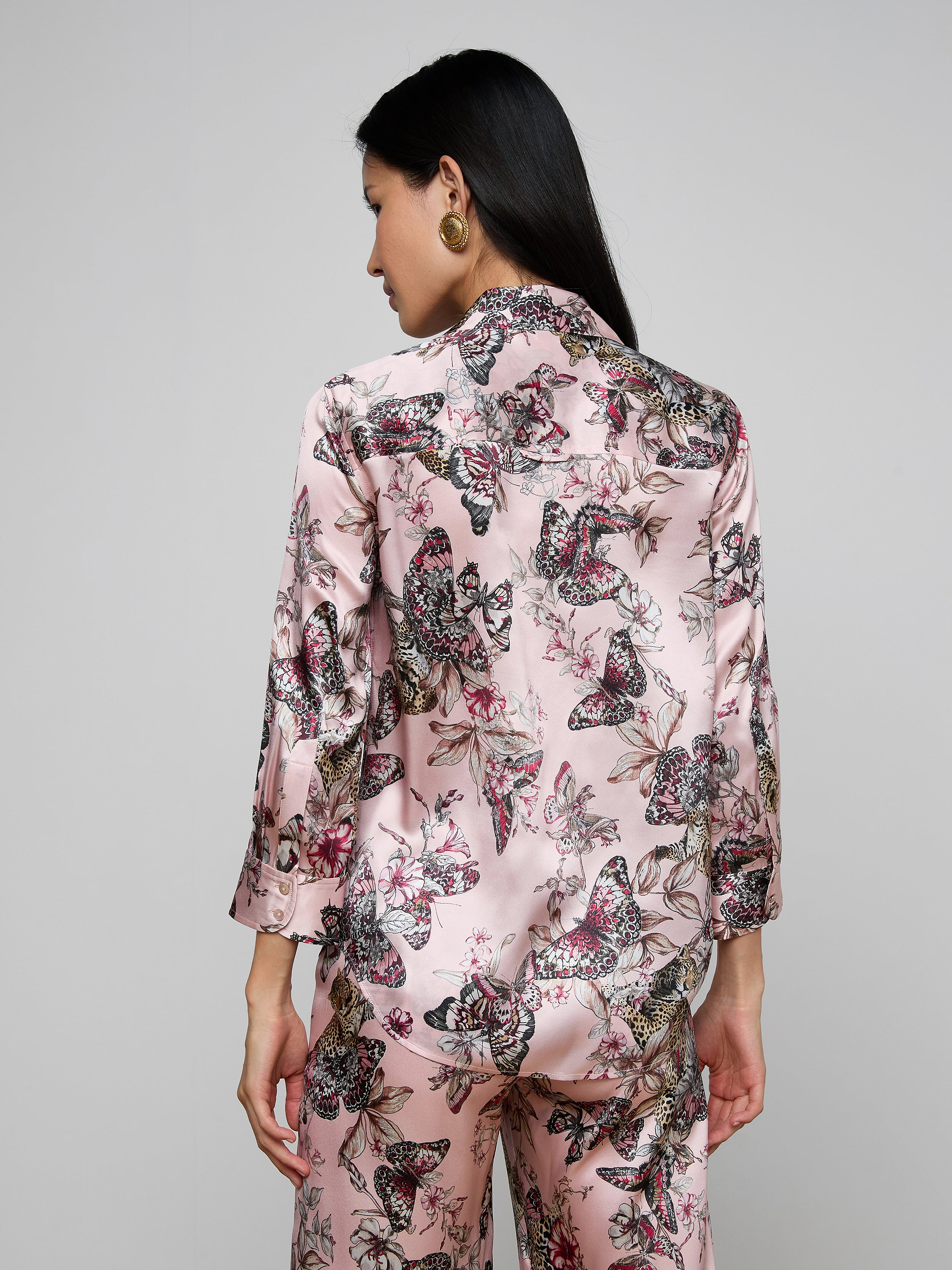 A woman with long dark hair, seen from the back, wears the Dani Silk Blouse featuring a pink butterfly jungle print, standing against a plain light background.