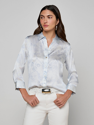A woman wears the Dani Silk Blouse, a light blue button-down with ivory and blue lace print, styled with white pants and a silver belt. She stands against a grey background, looking to the side with her hands in her pockets.