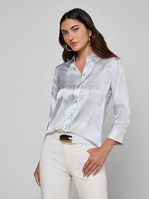 A woman with long brown hair wears the Dani Silk Blouse in ivory and blue lace print, paired with white pants, standing with one hand in her pocket against a plain grey background.