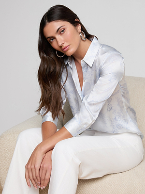 A woman with long brown hair, wearing an ivory and blue Dani Silk Blouse and white pants, sits calmly on a beige chair, hands on her knees, looking at the camera.